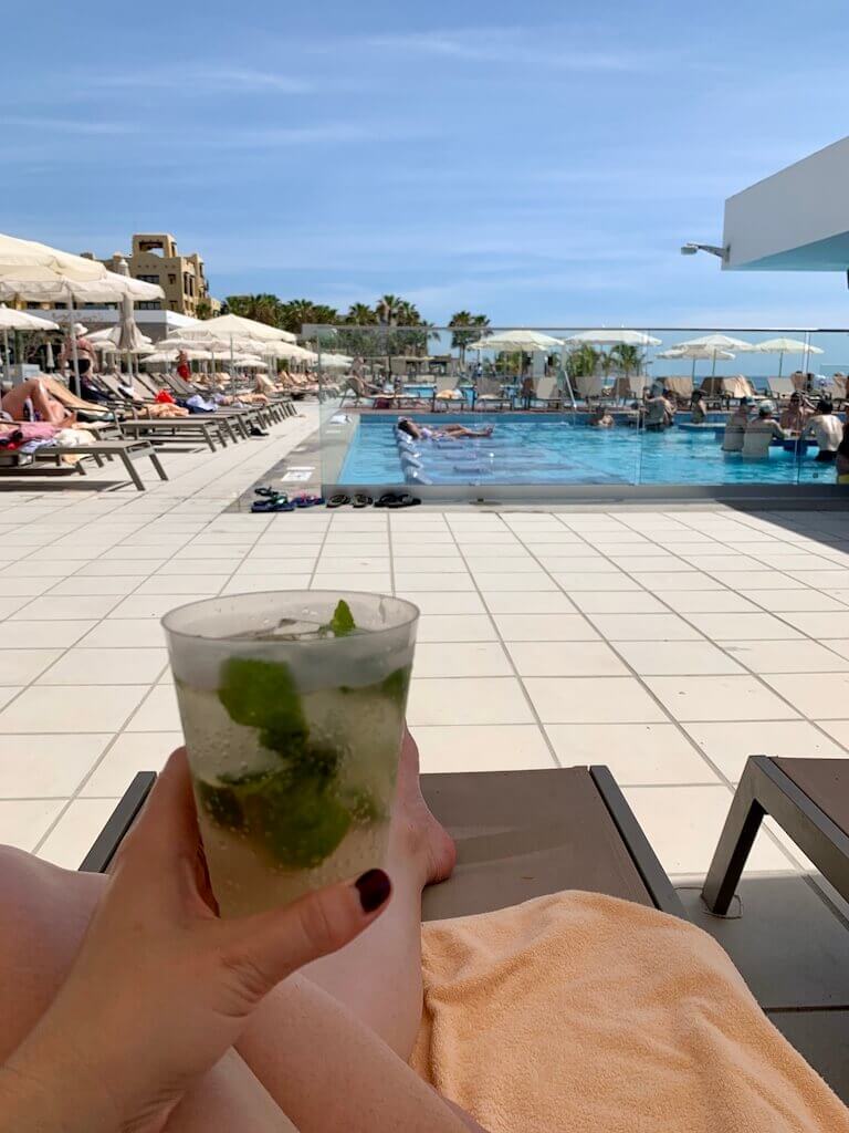 a woman holds a mojito in the foreground with a busy resort pool in the distance