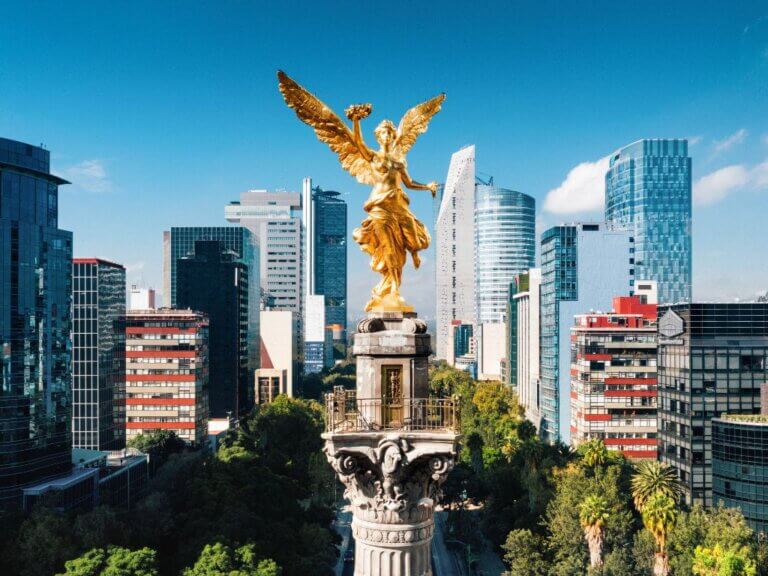 Golden Angel of Independence monument towers in the foreground with lush greenery, against a backdrop of modern skyscrapers under a clear blue sky in Mexico City, symbolizing the city's rich history and urban growth.