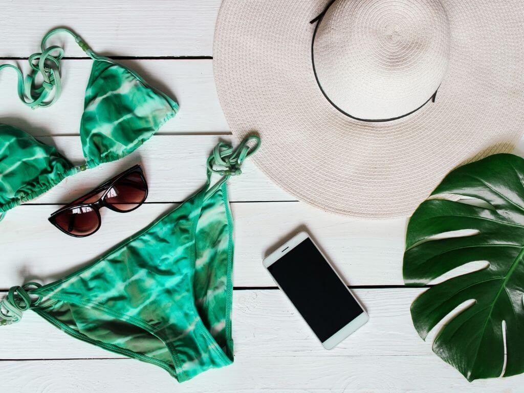 flatlay image of triangle string bikini with cell phone, sunglasses, and a sun hat