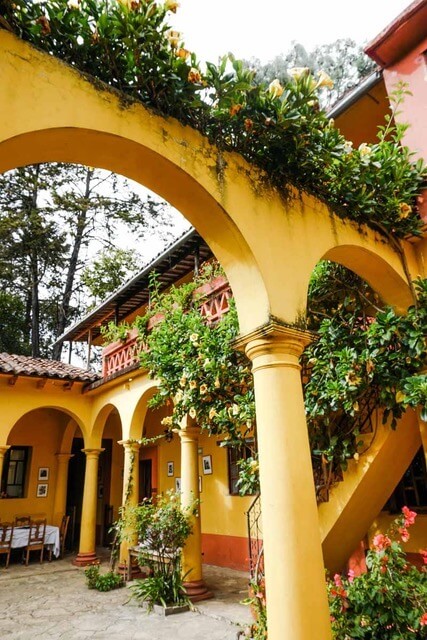 The courtyard of a colonial building features an abundance of potted plants and the architecture incorporates posts and arches, painted a warm shade of yellow