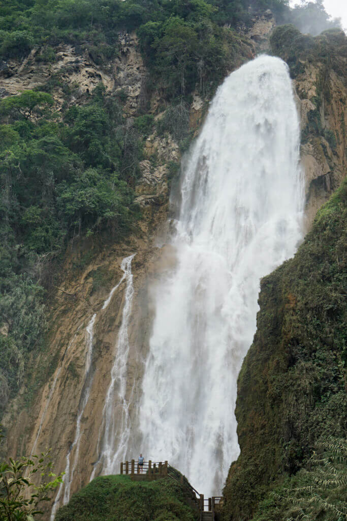 A traveler stands on a lookout point gazing up at the El Chiflon waterfalls in Chiapas, Mexico. The falls thunder down the cliff in the distance.