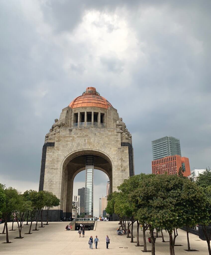 The Monument to the Revolution in Mexico City, a massive stone arch with a copper dome, stands against a dramatic sky, symbolizing the city's storied past.