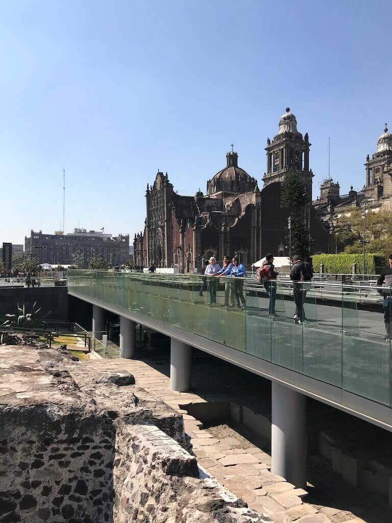 Visitors explore the glass-floored walkway above the ancient ruins at Templo Mayor with the Metropolitan Cathedral's ornate architecture in the distance, showcasing Mexico City's layered history.