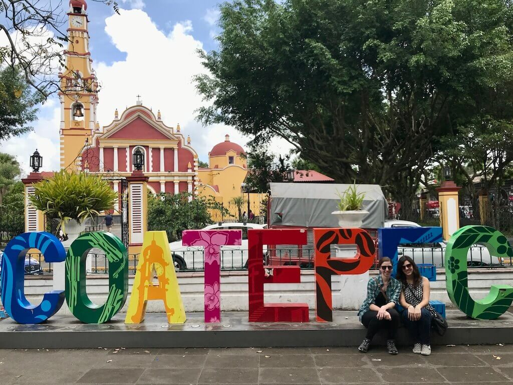 two girls sit in front of a colorful letter sculpture that reads Coatepec. There are leafy green trees in the background and a yellow and red cathedral