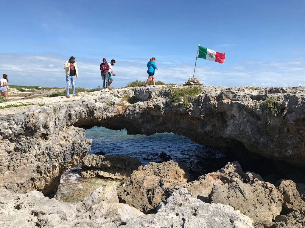 Tourst walk over a natural stone archway with ocean waves underneath at the El Mirador lookout on Cozumel. The Mexican flag flies in the distance,