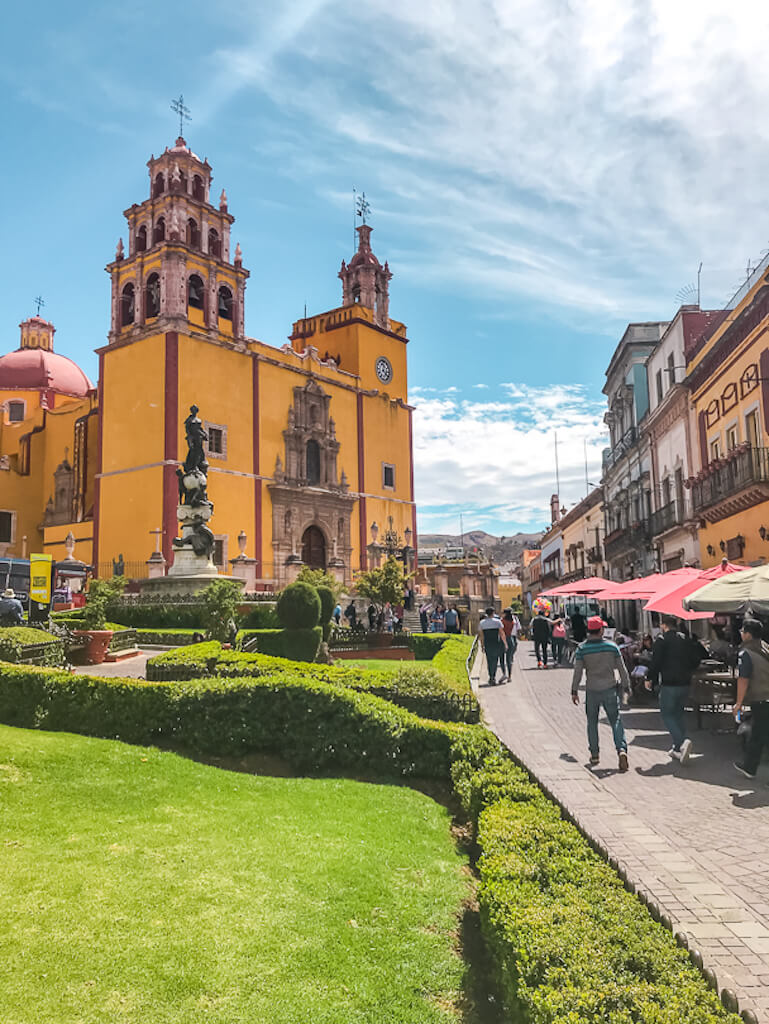 looking at Guanajuato's main cathedral with a patch of grass in the foreground and patio cafes stretching along the right side of the street