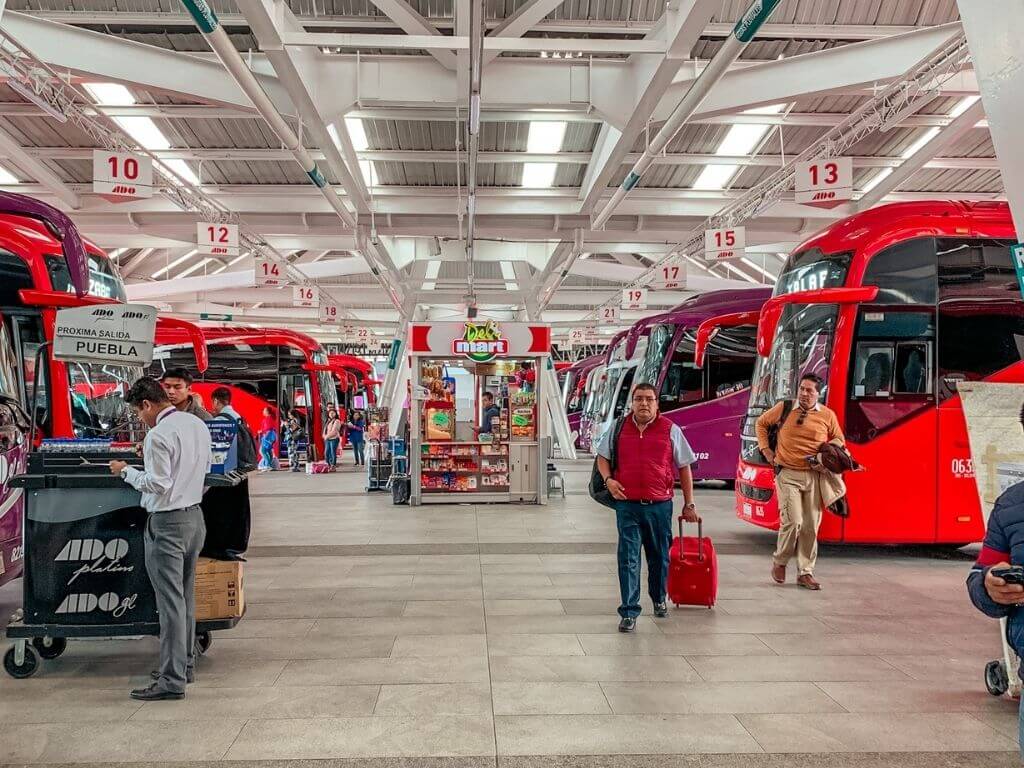 Mexico bus station with ADO buses lined up in bays on either side of a walkway. Travelers pulling suitcases approach the buses