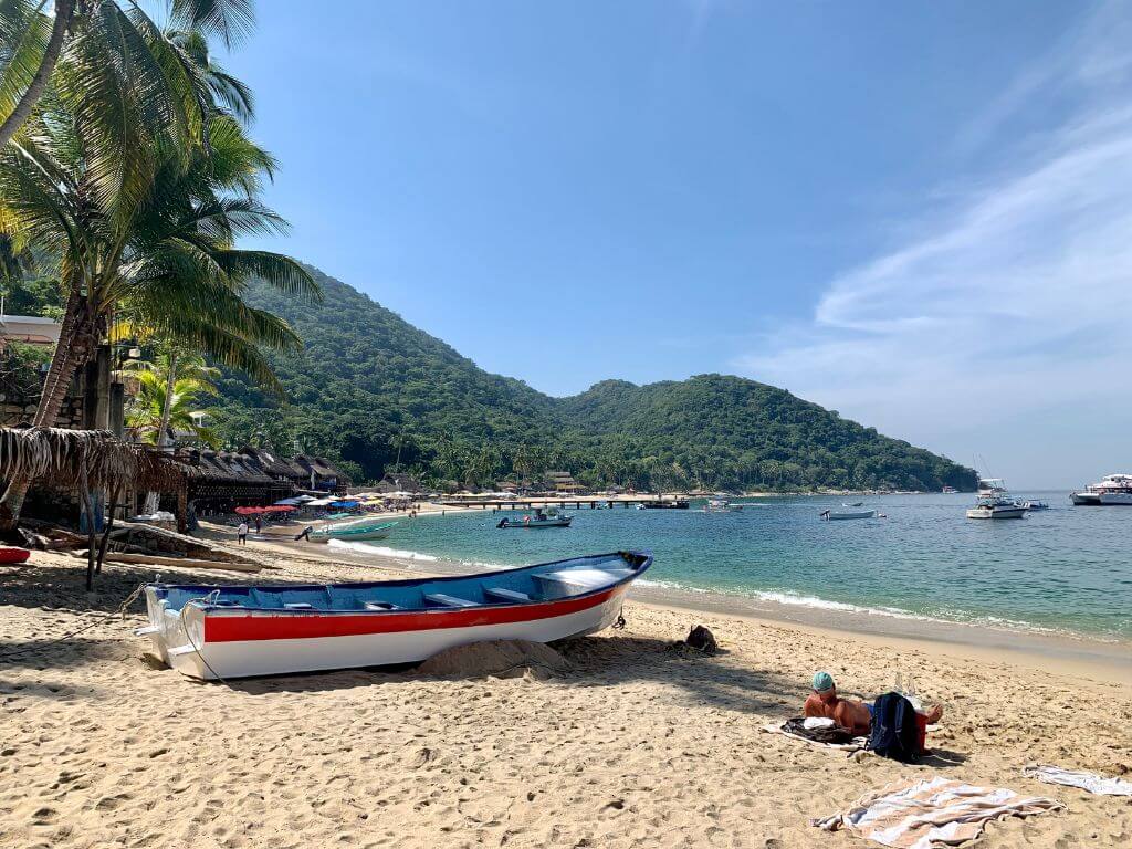 a panga boat and a sunbathing tourist sit in the sand in the foreground on Playa Las Animas with the bay and the green mountains in the distance