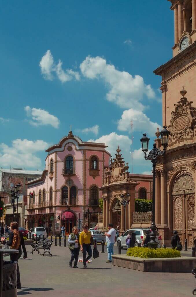 Scene of a busy street in Leon, Guanajuato with pedestrians walking through a square lined with colonial buildings while cars circle in the background