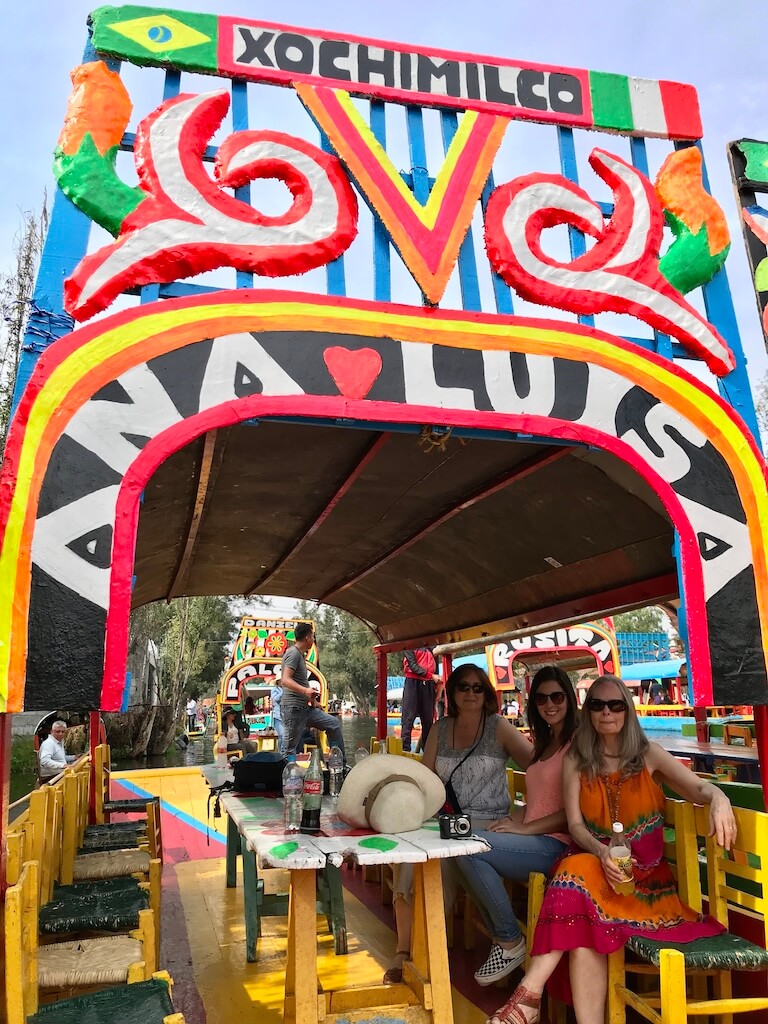 Three smiling women sit at a table in a trajinera with the name Ana Luisa