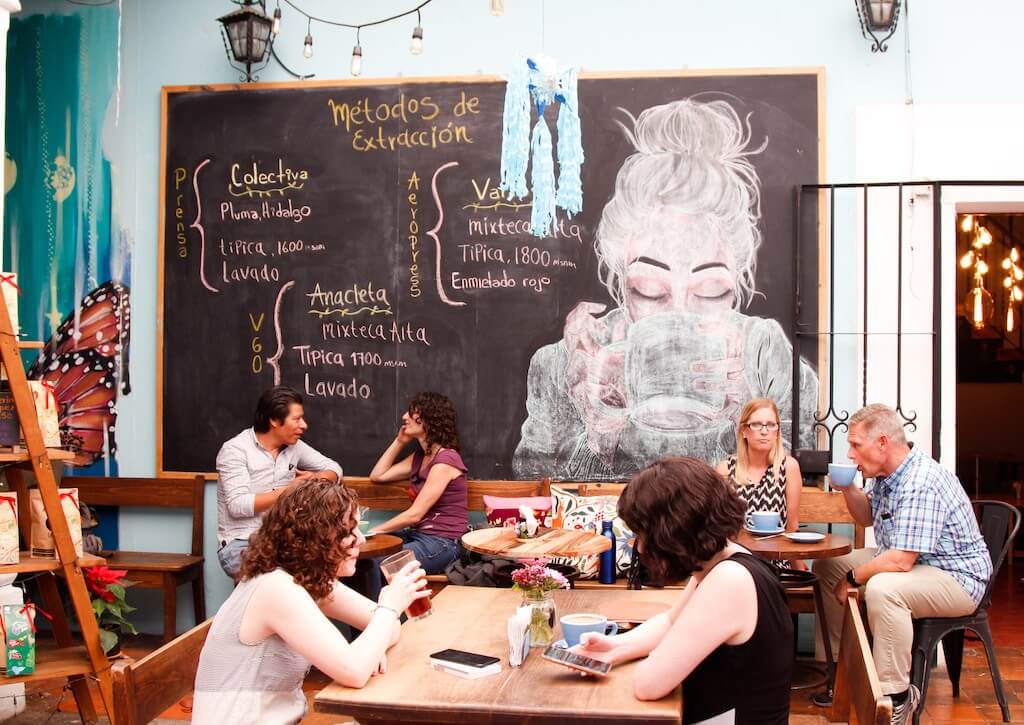 A cozy café in Oaxaca with patrons engaging in conversation, and a chalkboard displaying coffee extraction methods, representative of the inviting social scene in Oaxaca.