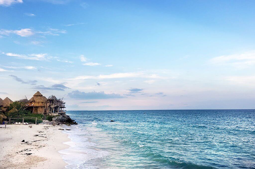 landscape view of a Riviera Maya beach at sunset. A thatched roof hotel lingers in the background and an empty beach and ocean make up the foreground