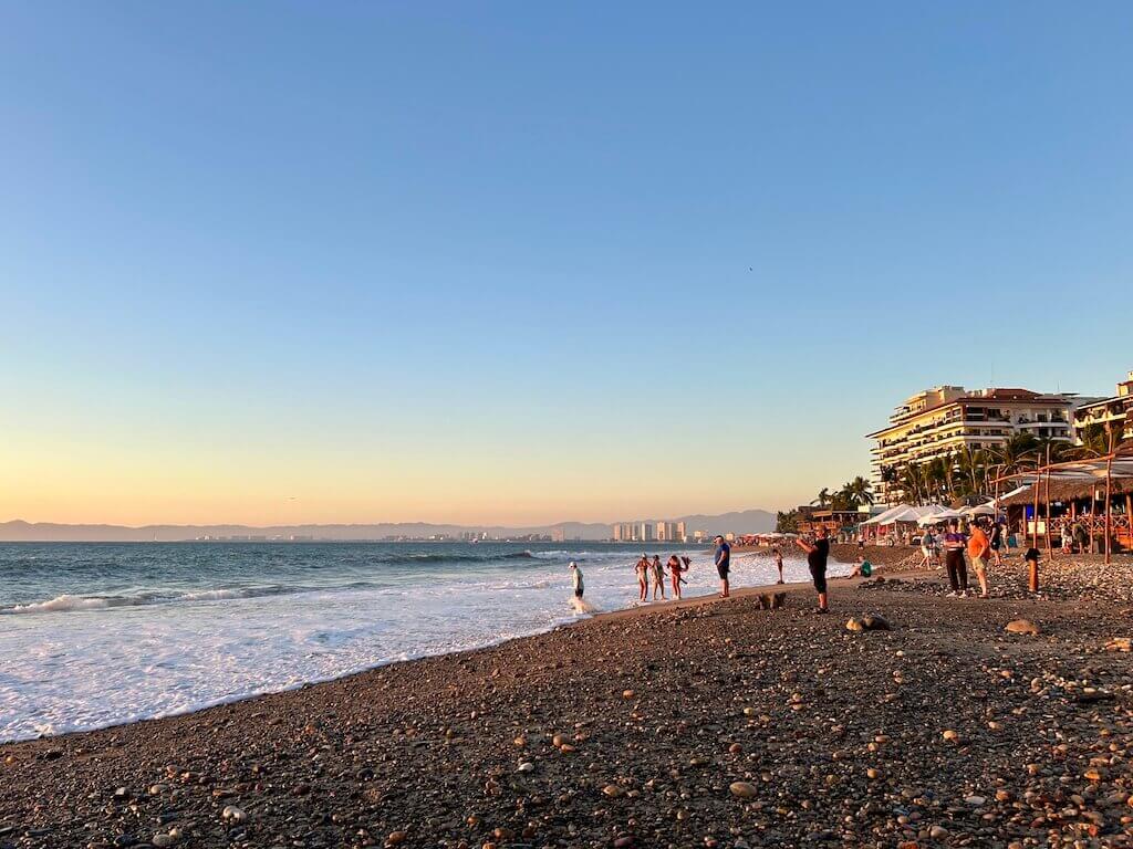 beachgoers congregate on playa camarones in Puerto Vallarta to capture photos of the sunset. looking north along the beach with the ocean lapping the shore on the left side of the image