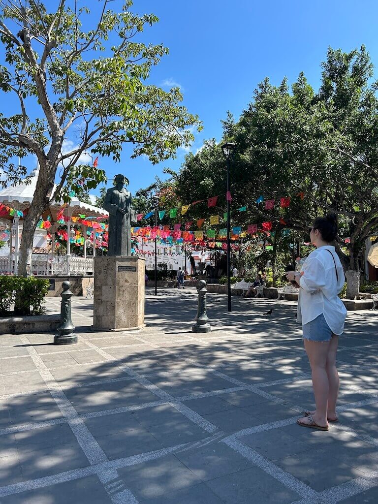 A woman admires a statue of a historic figure in Puerto Vallarta's plaza principal. Colorful papel picado is strung overhead. 