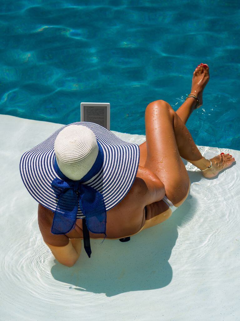 looking over the shoulder of a woman reading an ereader while laying partially in a swimming pool