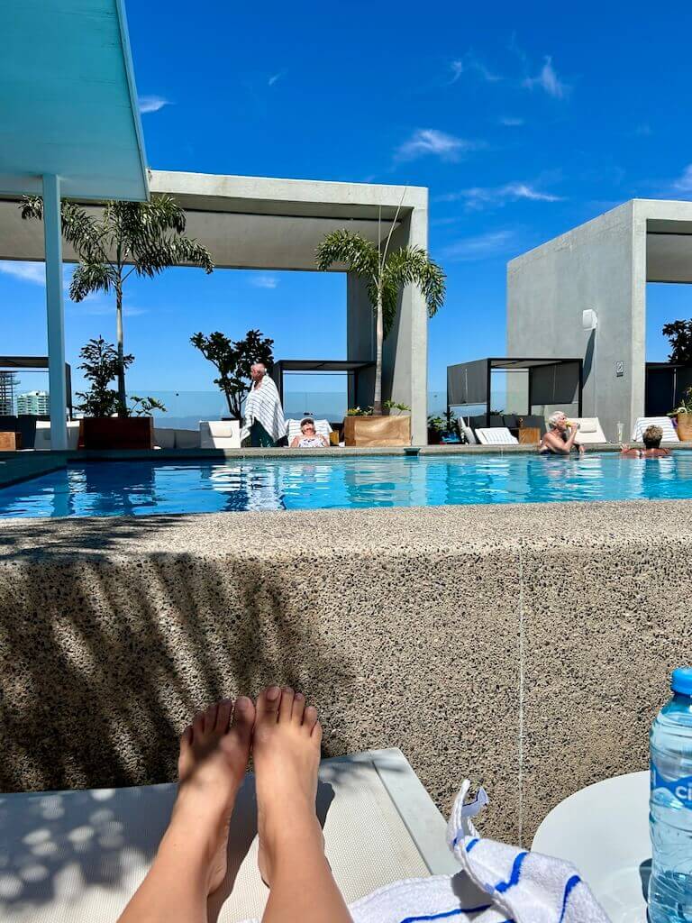 a woman's feet on a sun lounger looking toward a rooftop pool with ocean views in the distance