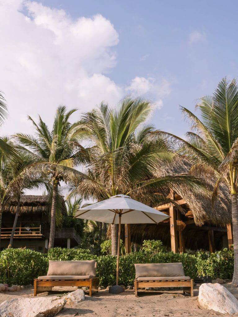 a pair of empty sun loungers with an umbrella between them on a sandy beach in Tulum, with palm trees in the background