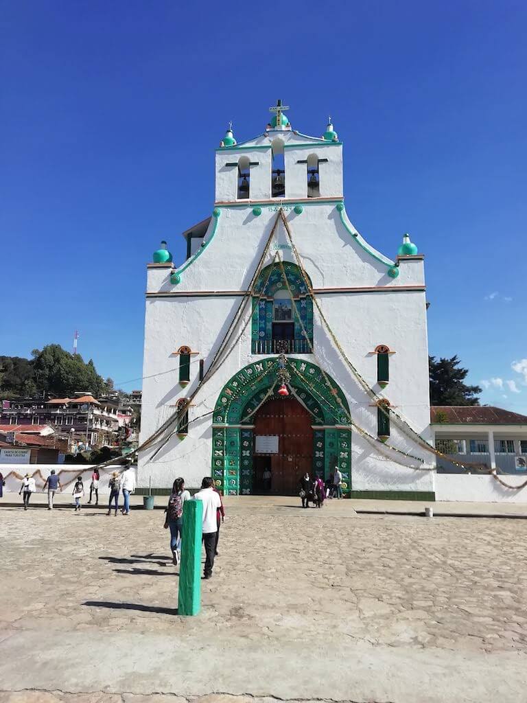 Folks approach the church of San Juan Chamula in Chiapas. The building is white with green trim, and an ornate green arched entrance with a heavy wood door. 