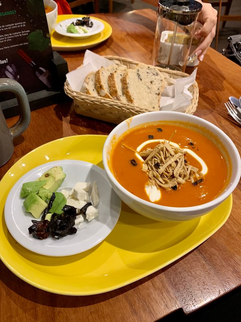 a bowl of tortilla soup with a plate of toppings, including avocado, dried chiles, and cheese, and a basket of fresh bread