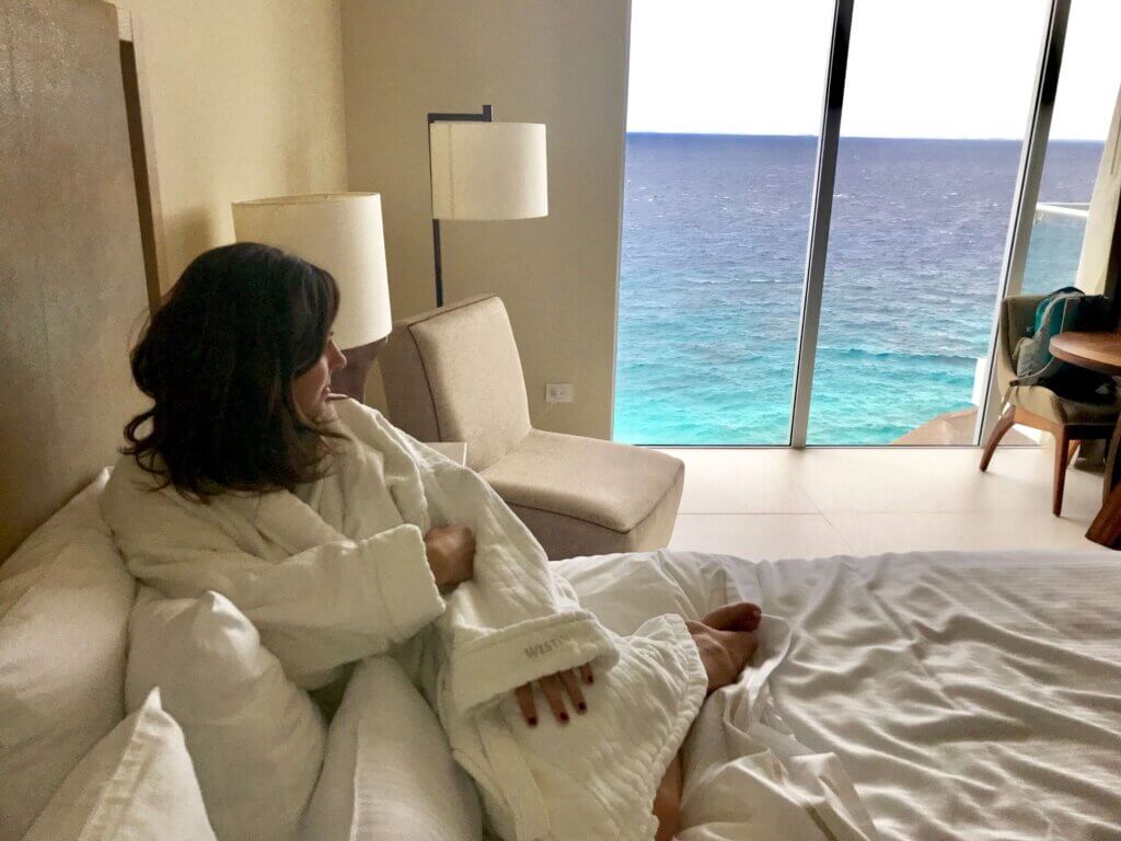 Janine wearing a fluffy white robe and looking out at the ocean view from a hotel room on Cozumel. 