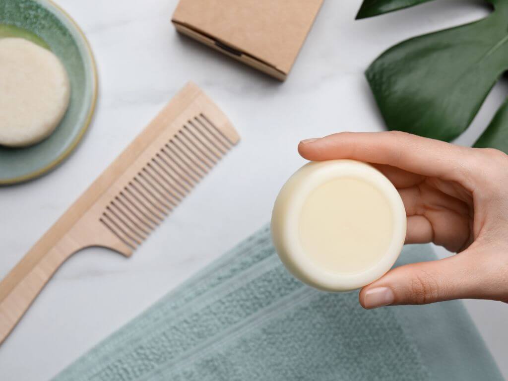 flat lay of someone holding a solid shampoo bar between their fingers with a comb and towel in the background