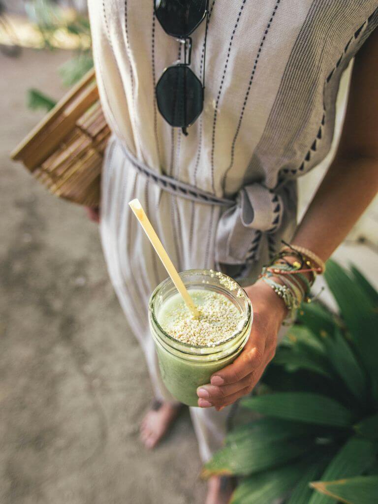 a woman in a gauzy white dress with black embroidery holds a green matcha drink in a mason jar. 