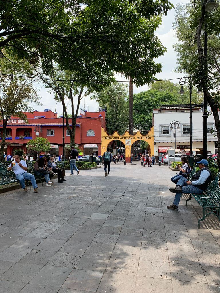 park in Coyoacan, Mexico City with the Artisan Market in the distance. people sit on park benches on either side of a walkway