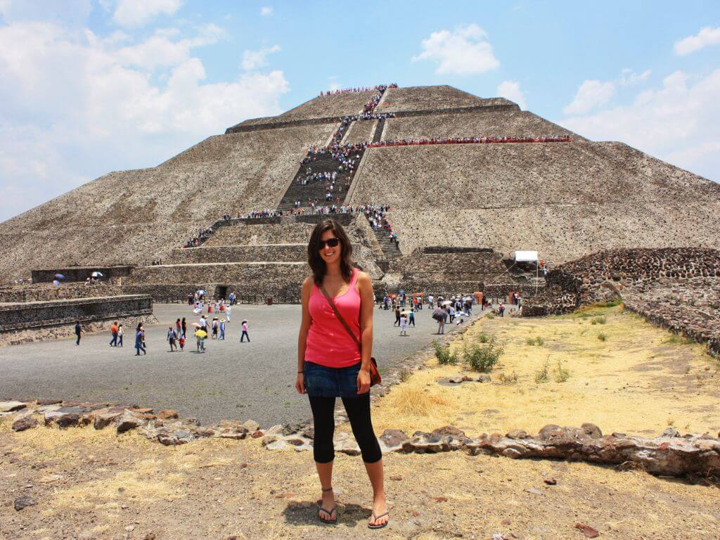 a woman wearing a pink tank top and a denim skirt stands in front of the pyramid of the sun at the Teotihuacan archaeological site 