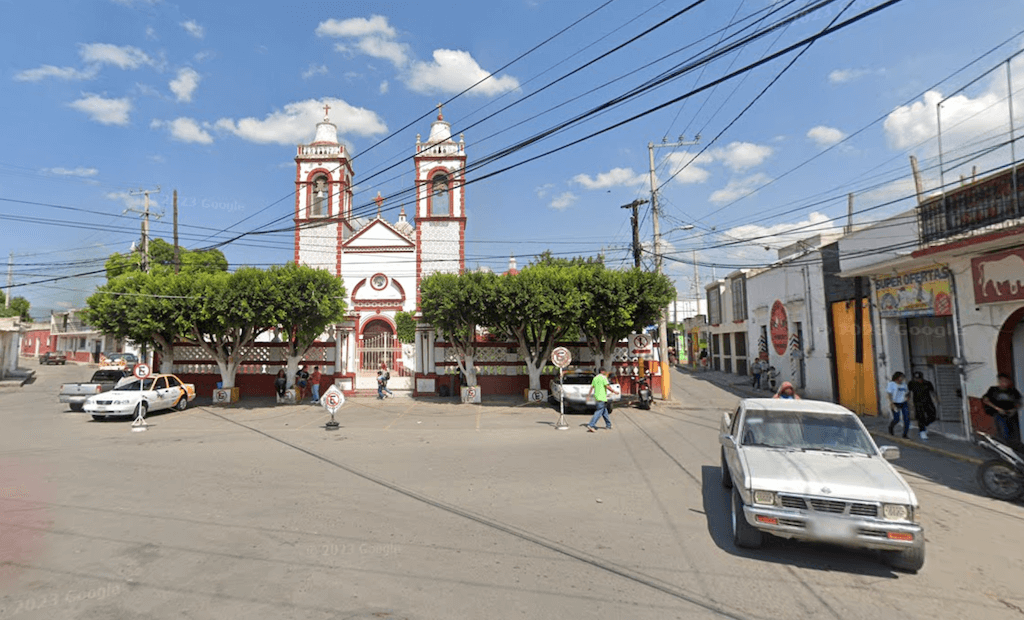 San Antonio Church in Ixmiquilpan bordered by green trees, with a wide parking area in front and cars circling around