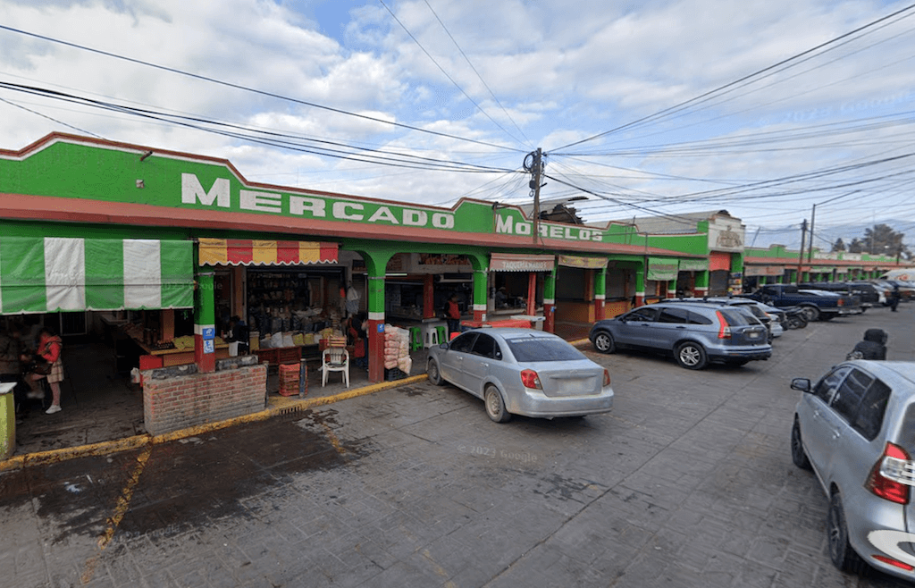 exterior of mexican marketplace; a green building that reads Mercado Morelos in white lettering above the market stalls. There are parked cars in the foreground.